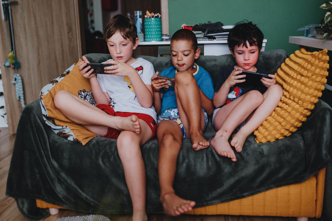 Three boys engrossed in mobile games while sitting on a couch at home, reflecting modern childhood leisure.